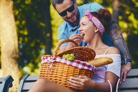 Tattooed bearded male and redhead female are having a picnic on a bench in a park.の写真素材