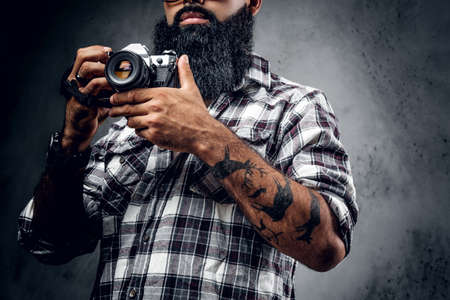 A bearded hipster amateur photographer with tattoos on arms, dressed in a fleece shirt holds compact DSLR camera over grey background.の写真素材