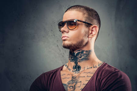 Handsome, athletic male in sunglasses with tattoos on his arms and neck posing over grey artistic background in a studio.の写真素材