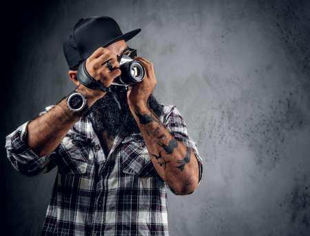 A bearded hipster amateur photographer with tattoos on arms, dressed in a fleece shirt holds compact DSLR camera over grey background.の写真素材