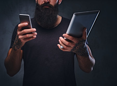 The tattooed, bearded hipster male holds tablet PC and smartphone on grey background.