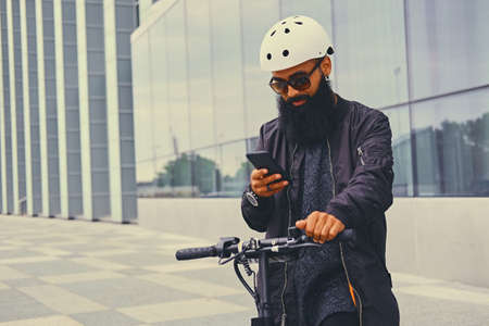 Shaved head tattooed, bearded male using a smart phone over modern building background after the electric scooter ride.の写真素材