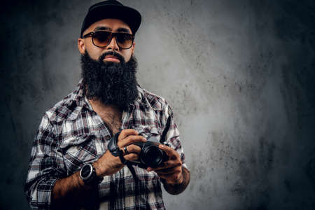 A bearded hipster amateur photographer with tattoos on arms, dressed in a fleece shirt holds compact DSLR camera over grey background.の写真素材