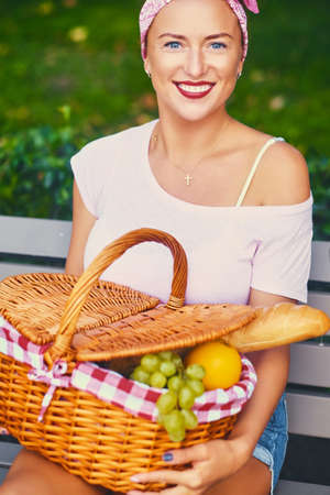 Positive redhead female sits on a bench with a picnic basket full of fruits, bread and wine.の写真素材