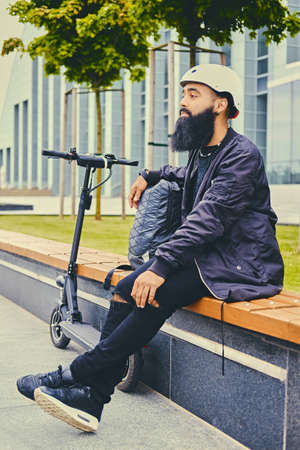 Stylish bearded male in sunglasses relaxing on a bench after riding by electric scooter in downtown.の写真素材