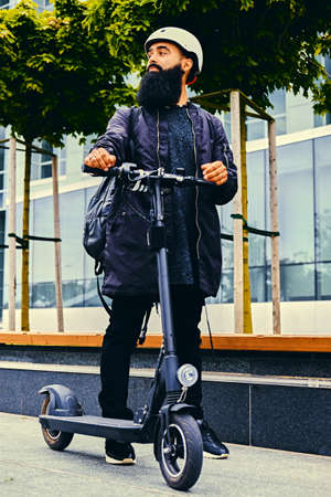 Stylish bearded male in sunglasses posing on electric scooter in over modern building background.