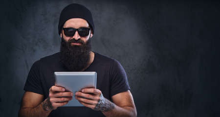 Bearded brutal male in a hat and sunglasses holds tablet PC over grey background.の写真素材