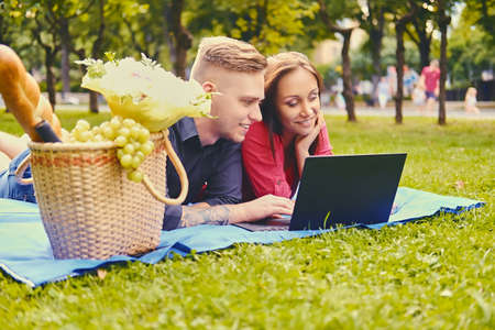 Attractive couple lying on a blanket on a lawn and is using a laptop on a picnic.の写真素材