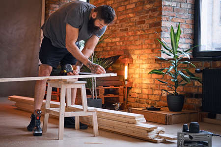 The full body image of bearded tattooed carpenter drilling a hole in a board in a room with loft interior.の写真素材