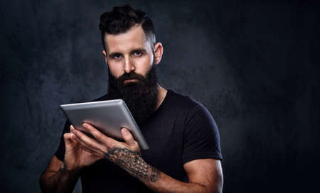 Bearded male with tattooed arms holds silver tablet PC over grey background.