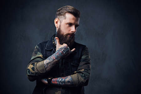 Portrait of bearded tattooed hipster male dressed in a military jacket and black waistcoat over grey background.の写真素材