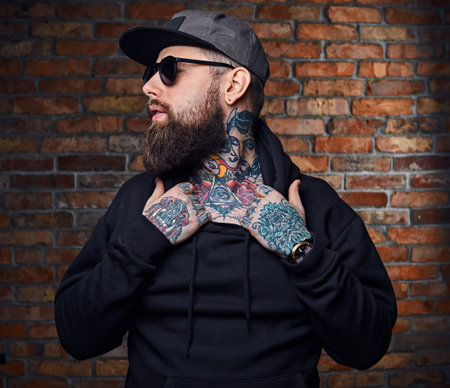 Modern bearded hipster male with tattoos on his arms, dressed in a military jacket posing over...
