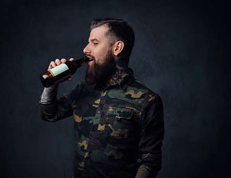A tattooed, bearded hipster male dressed in military shirt, drinks craft beer from a bottle.の写真素材