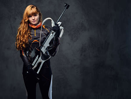 Studio portrait of a redhead female Biathlon champion holds a gun over grey background.