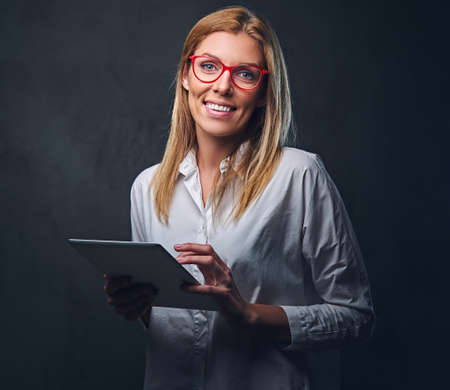 A blond female dressed in a white shirt and eyeglasses holds tablet PC on grey background.