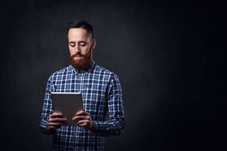 Redhead bearded male dressed in a blue fleece shirt holds tablet PC on grey background.の写真素材