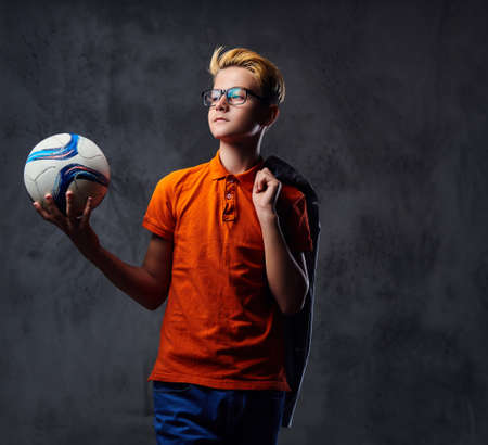 A blond teenager boy dressed in a jeans and orange t shirt holds a soccer ball over grey background.の写真素材