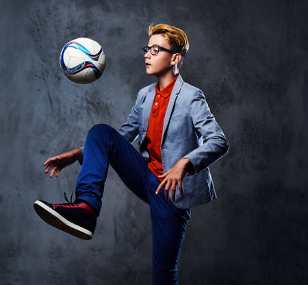 A blond teenager boy dressed in a jeans plays with a soccer ball over grey background.の写真素材