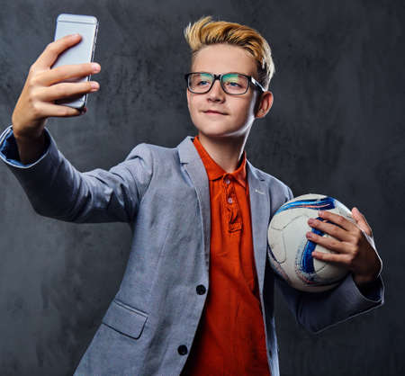 Blond teenager boy in eyeglasses holds soccer ball and making selfie on a smartphone.