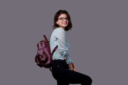 Positive young traveler female with leather backpack isolated on grey background.