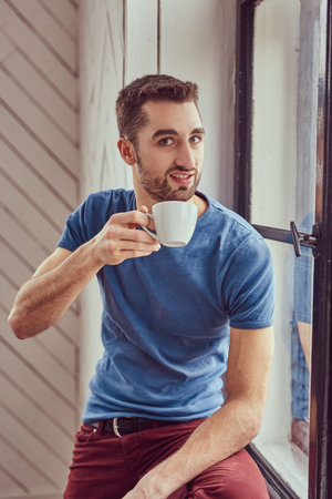 Young caucasian student drinks morning coffee sitting on a windowの写真素材