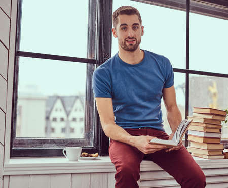 Young caucasian student reading a book sitting on a window sill.の写真素材