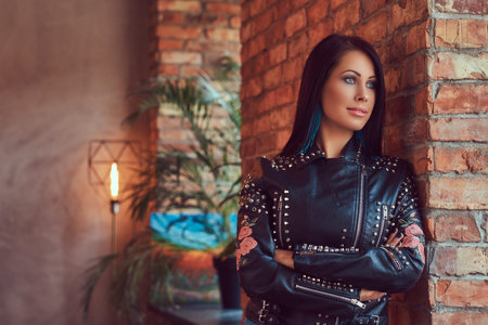 Sexy sensual brunette posing in stylish leather jacket and jeans leaning against a brick wall.
