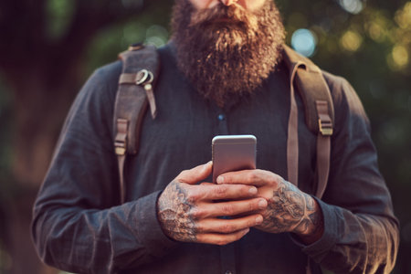 A handsome hipster traveler with a stylish beard and tattoo on his arms dressed in casual clothes and hat, using the phone.の写真素材