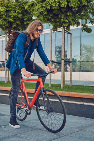 A handsome bearded male with long hair in stylish clothes and sunglasses with a backpack, riding a bicycle on the street, outdoors.の写真素材