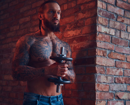 A handsome tattoed shirtless male with a stylish haircut and beard, doing exercise with a dumbbell,...