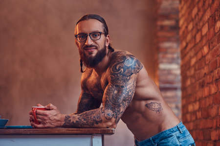 A handsome bearded tattoed male with a stylish haircut and muscular body, drinks coffee, leaning on a table.の写真素材