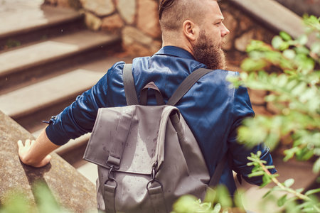 Back view portrait of a bearded male with a haircut dressed in casual clothes with a backpack, sitting in a city parkの写真素材