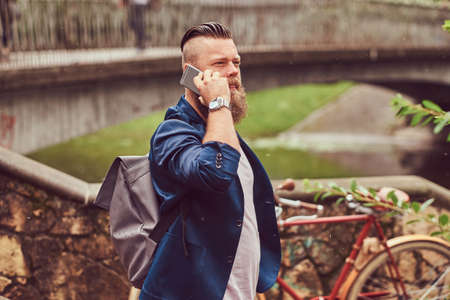 Portrait of a bearded male with a haircut dressed in casual clothes with a backpack, standing in a park, talking by phone.の写真素材
