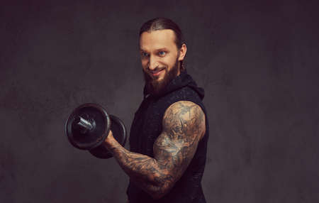 Portrait of a muscular bearded tattoed male with a stylish haircut wearing black sportswear, doing exercise with a dumbbell, isolated on a dark background.の写真素材
