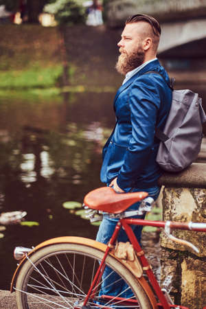 Bearded male with a stylish haircut dressed in casual clothes with a backpack, standing with a retro bicycle near the river in a city park.の写真素材