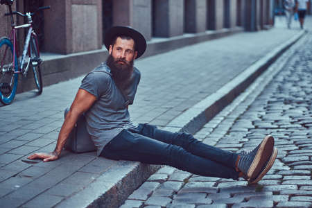 A handsome hipster traveler with a stylish beard and tattoo on his arms dressed in casual clothes with a bag, sits on the sidewalk, resting after a bike ride.の写真素材