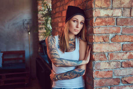 A tattoed sexy blonde in a t-shirt and a hat posing against a brick wall.の写真素材