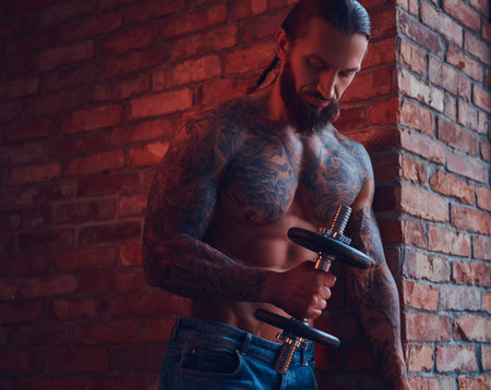 A handsome tattoed shirtless male with a stylish haircut and beard, doing exercise with a dumbbell, standing against a brick wall in a room with a loft interior.の写真素材