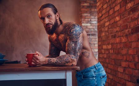 A handsome bearded tattoed male with a stylish haircut and muscular body, drinks coffee, leaning on a table.の写真素材