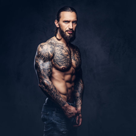 Close-up portrait of a muscular naked bearded tattoed male with a stylish haircut, isolated on a dark background.の写真素材
