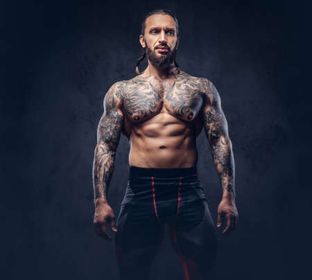 Portrait of a muscular naked bearded tattoed male with a stylish haircut in a sports trousers, isolated on a dark background.の写真素材