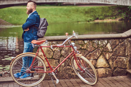 Bearded male with a stylish haircut dressed in casual clothes with a backpack, standing with a retro bicycle near the river in a city park.の写真素材