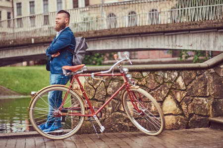 Bearded male with a stylish haircut dressed in casual clothes with a backpack, standing with a retro bicycle near the river in a city park.の写真素材