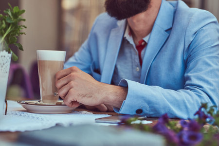 Portrait of a fashionable bearded male with a stylish haircut, holds a glass of a cappuccino, sitting in a cafe outdoors.の写真素材