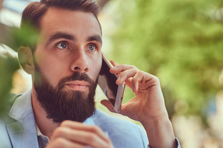 Close-up portrait of a fashionable bearded male with a stylish haircut, speaking by phone, sitting in a cafe outdoors.の写真素材
