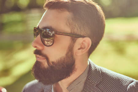 Close-up portrait of a bearded male with a stylish haircut, in a jacket and sunglasses, sitting on a bench in a park.の写真素材