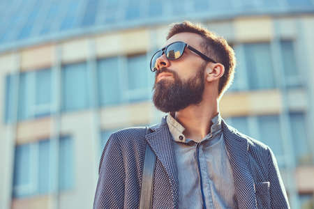 Close-up portrait of a bearded male wearing casual clothes and sunglasses, standing in a city against a skyscraper.の写真素材