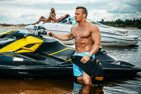 Handsome shirtless sportsman with a muscular body standing near jet ski on a seacoast.の写真素材