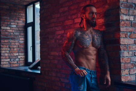 A handsome tattoed shirtless male with a stylish haircut and beard, standing against a brick wall in...