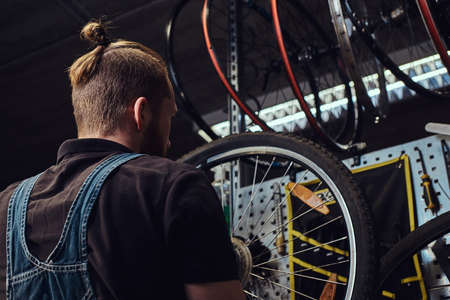 Handsome redhead male in a jeans coverall, working with a bicycle wheel in a repair shop. A worker removes the bicycle tire in a workshop.の写真素材